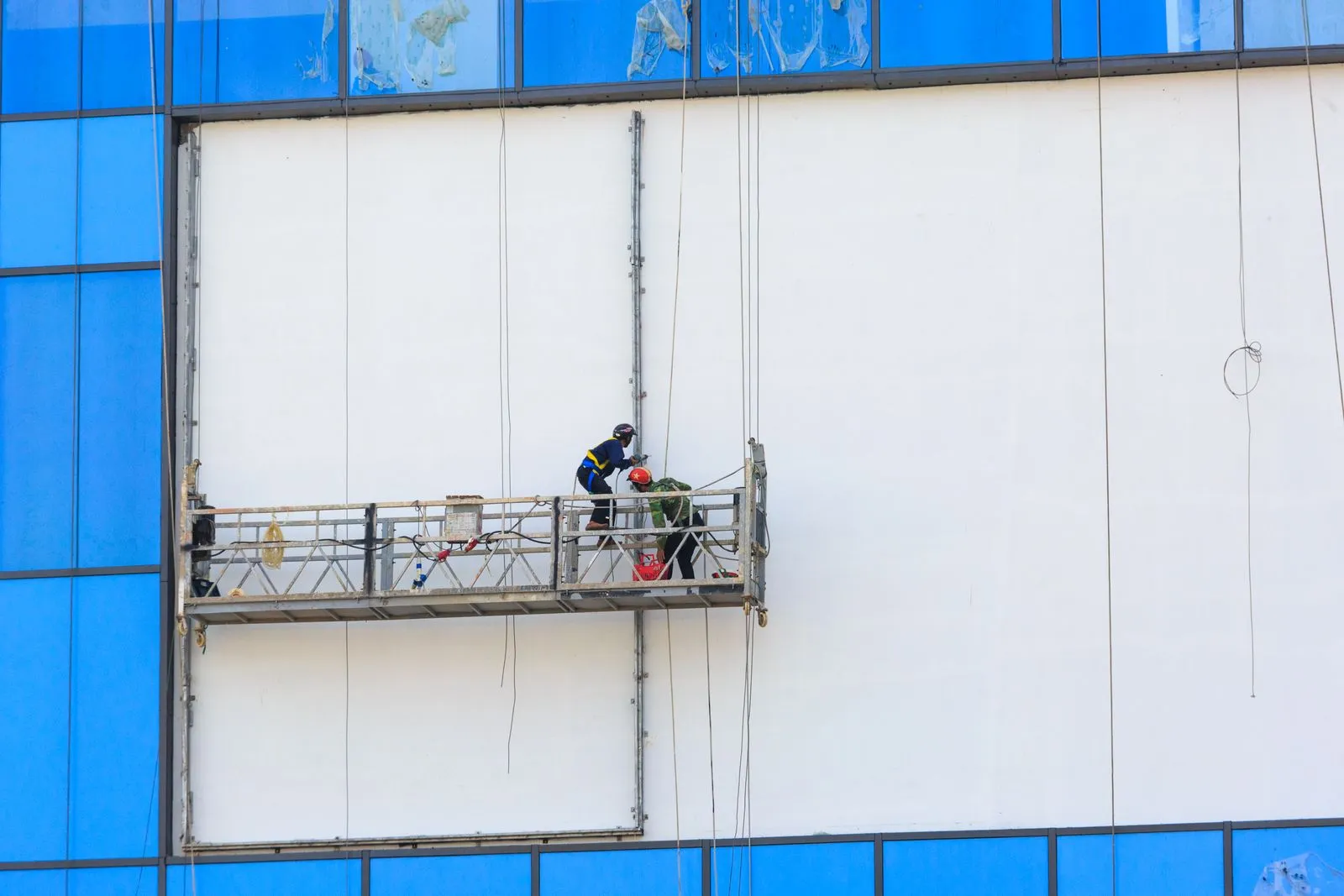 Grand pan de façade blanc entre des vitrages bleus, nacelle de chantier